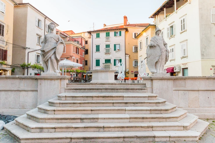 the First of May Square in Piran, with a stone cistern flanked by the statues representing Strength and Vigilance