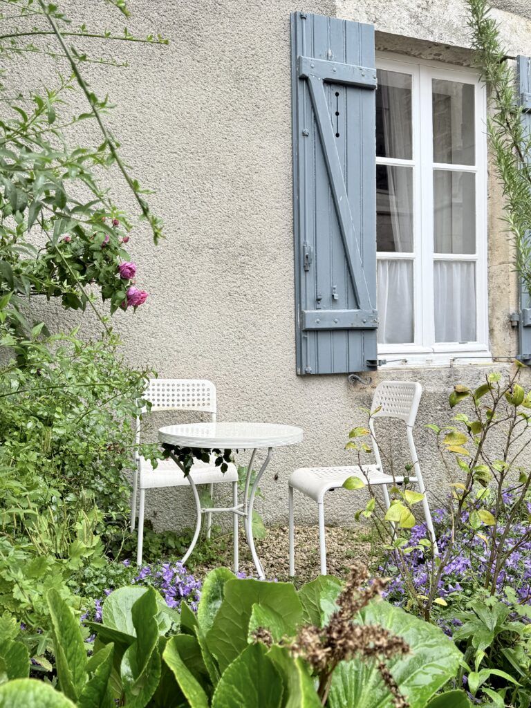 house with blue shutters and a cafe table and chair with purple flowers and roses